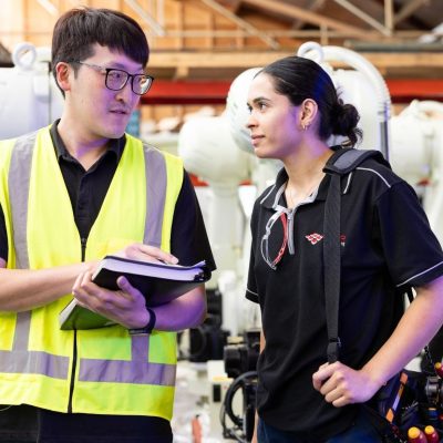 Two apprentices in hi-vis vests discussing notes inside a workshop.